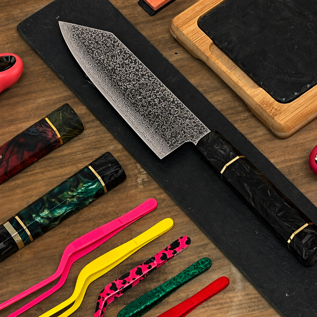 Cuchillo Bunka Damascus VG10 con hoja de acero Damascus y mango de fibra de carbono sobre tabla negra, vista cenital con utensilios de cocina